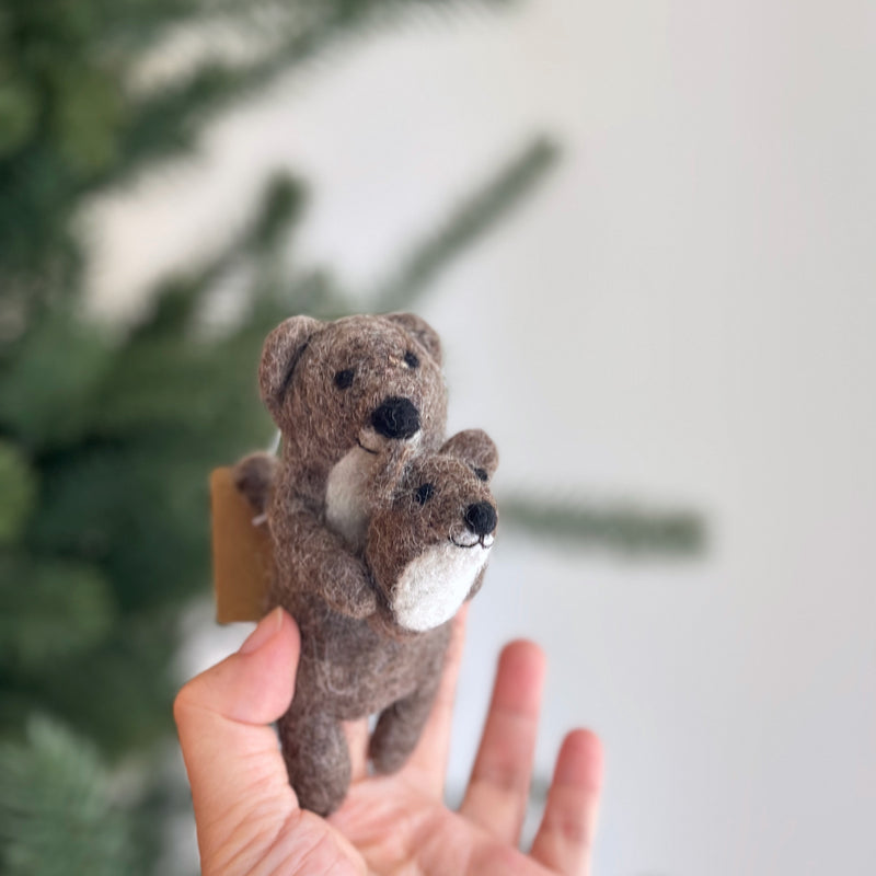A handmade felt American River Otter finger puppet, featuring a light brown mother otter cradling a smaller white baby otter with a brown face, is held by a fair-skinned hand. Both otters have black eyes and noses. This unique handcrafted felt puppet is set against a blurred Christmas pine tree and a light gray wall.