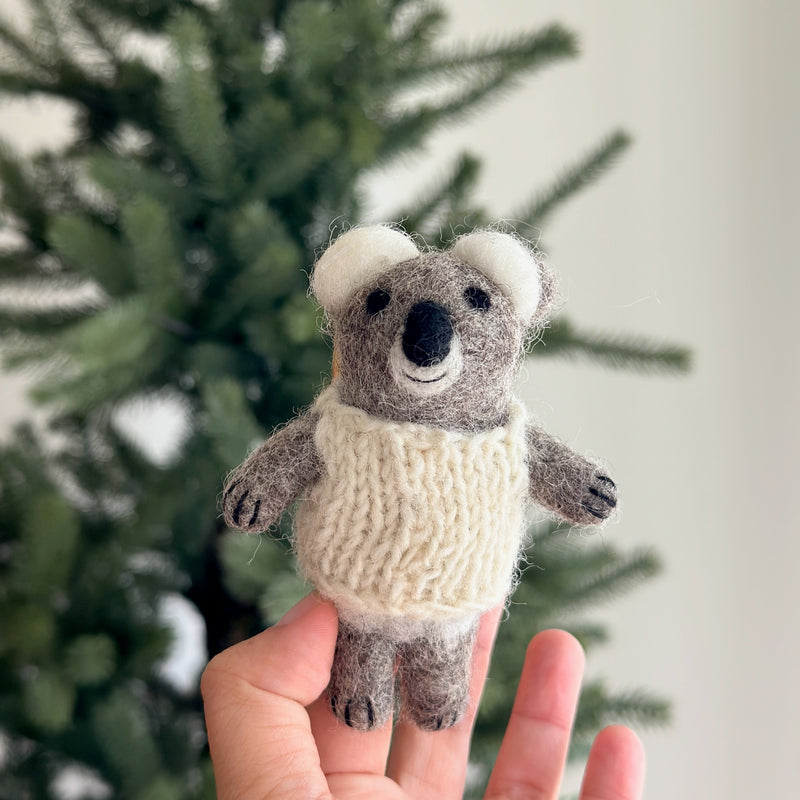 A cozy, handmade wool felt baby koala finger puppet is gently held by a female hand, set against a blurred, warm background suggesting a Christmas tree. This adorable koala puppet, crafted from soft gray wool felt, wears a white woven sweater and features small round white ears, a black nose, and tiny black claws. This unique, handcrafted felt toy is lovingly made in Nepal, reflecting its fair-trade origin and artisanal quality.