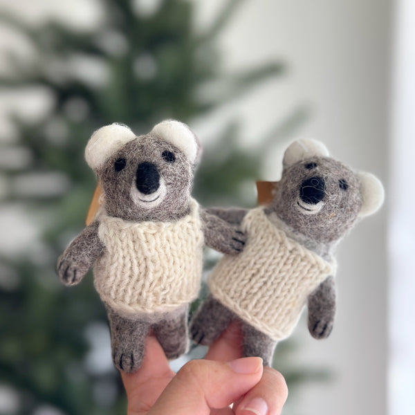 A meticulously handcrafted wool felt baby koala finger puppet is gently held in a hand, displayed against a softly blurred festive Christmas tree backdrop, evoking a playful and inviting mood. The charming gray koala puppet features soft gray fur, fluffy white ears, small black eyes, and a stitched smile, all adorned with a cozy hand-knitted white sweater.