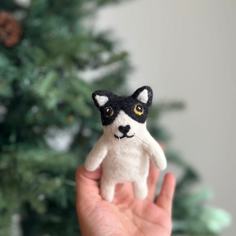 A handcrafted needle felted tuxedo cat finger puppet is held in a hand, appearing against a soft, blurred background that suggests a Christmas tree. This charming felt toy features a white body with distinctive black markings around its eyes and on its head, resembling a tuxedo pattern. It has round yellow eyes, a black heart-shaped nose, and delicate whiskers, showcasing its tactile appeal for imaginative play.