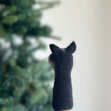A handcrafted wool felt turtle finger puppet is held up by a hand against a blurred Christmas tree background. This unique needle felted turtle puppet, made from 100% wool, showcases subtle textural variations. Its artisanal quality stands out vibrantly against the soft, out-of-focus green of the tree.