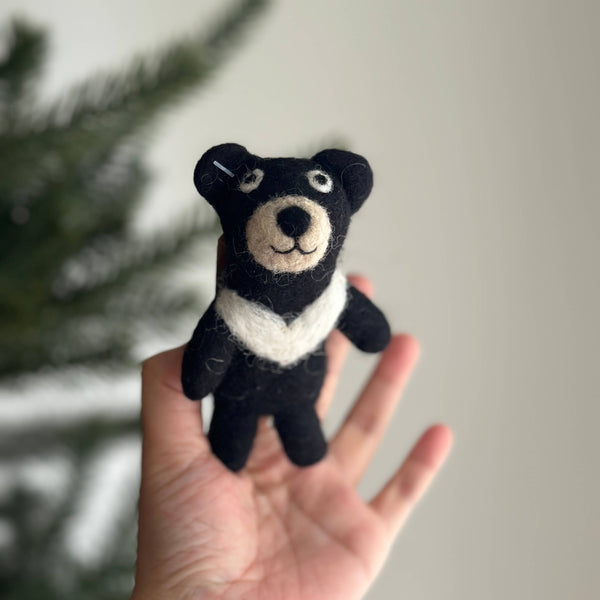 A handmade wool felt Taiwanese Black Bear finger puppet is presented held gently in a female hand, set against a softly blurred Christmas tree background. The bear features a soft, black body, a beige face, and a distinctive white crescent marking on its chest. With small, rounded ears and simple stitched facial features, this approximately 4-inch tall puppet embodies unique craftsmanship.