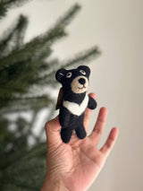 A handcrafted wool felt Taiwanese Black Bear finger puppet is held in a female hand against a softly blurred background. The black bear puppet features a distinctive white crescent on its chest, a beige face, and inner ears, with stitched black eyes and a smiling mouth. Its needle-felted wool gives it a soft, velvety texture, highlighting its unique design.