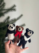A black bear felt finger puppet is playfully displayed in a female hand, set against a soft white background with a hint of Christmas tree branches, creating a festive mood. This handcrafted wool felt black bear finger puppet features a distinctive white felt crescent on its chest, showcasing its detailed craftsmanship and soft texture, ideal for imaginative play.