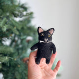 A handmade black cat felt finger puppet is presented by a female hand, set against a softly blurred Christmas tree backdrop. This charming felt toy for kids features captivating yellow big eyes, a unique love-shaped gray nose, and delicate whiskers. The black felt cat's inner ears are a soft pink, adding a playful touch to this handcrafted felt puppet.