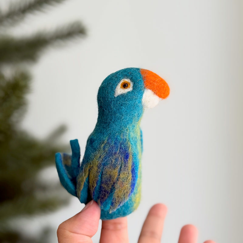 A vibrant needle felted blue parrot finger puppet is held upright on a finger against a soft white backdrop with a blurred Christmas pine tree. This handcrafted wool felt toy features striking blue, yellow, and white textures, highlighted by a contrasting orange beak. Its 3D design makes it an engaging prop for storytelling and imaginative kids' play.