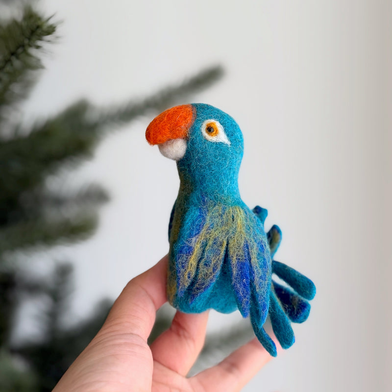 A handcrafted needle felted blue parrot felt finger puppet is gently presented by a hand, set against a softly blurred Christmas tree backdrop. This unique 3D designed wool felt finger puppet features shades of blue with yellow accents, an orange beak, and expressive white and black eyes, highlighting its detailed craftsmanship.