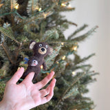 A needle-felted brown bear finger puppet is held up by a fair-skinned hand against a festive, blurred Christmas tree backdrop. This handcrafted brown bear puppet features light beige accents on its ears and face. It proudly holds a white felt fish with blue fins and a red smile. This unique, fair-trade felt toy, crafted from wool, evokes a sense of festive imagination.