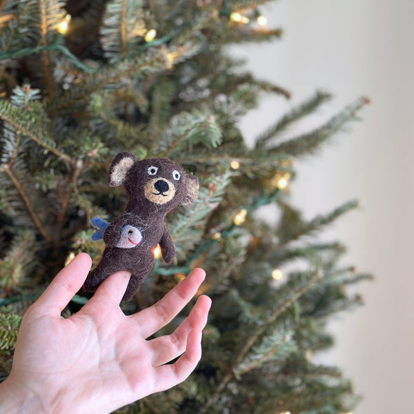 A needle-felted brown bear finger puppet is held up by a fair-skinned hand against a festive, blurred Christmas tree backdrop. This handcrafted brown bear puppet features light beige accents on its ears and face. It proudly holds a white felt fish with blue fins and a red smile. This unique, fair-trade felt toy, crafted from wool, evokes a sense of festive imagination.