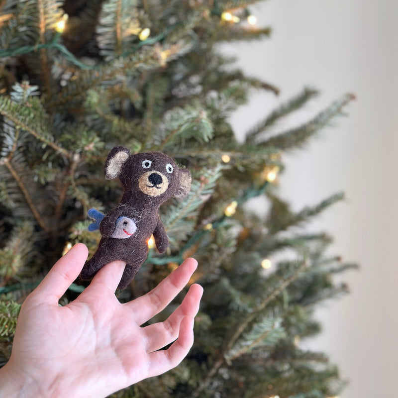 A needle-felted brown bear finger puppet is held up by a fair-skinned hand against a festive, blurred Christmas tree backdrop. This handcrafted brown bear puppet features light beige accents on its ears and face. It proudly holds a white felt fish with blue fins and a red smile. This unique, fair-trade felt toy, crafted from wool, evokes a sense of festive imagination.
