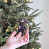 A handmade brown bear wool felt finger puppet, holding a fish, is delicately held on a fair-skinned female finger, set against a blurred, festive Christmas tree backdrop. This charming felted finger puppet features a beige snout and round ears, highlighting its handcrafted charm and suitability for engaging play.