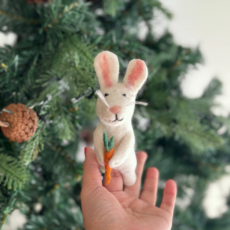 A handmade felt bunny finger puppet holding a carrot is gently cradled in a female hand, with a blurred Christmas tree visible in the background. The charming white bunny puppet features long ears with pink inner details, tiny black eyes, and whiskers, holding a small orange and green felt carrot, emphasizing its soft, imaginative design.