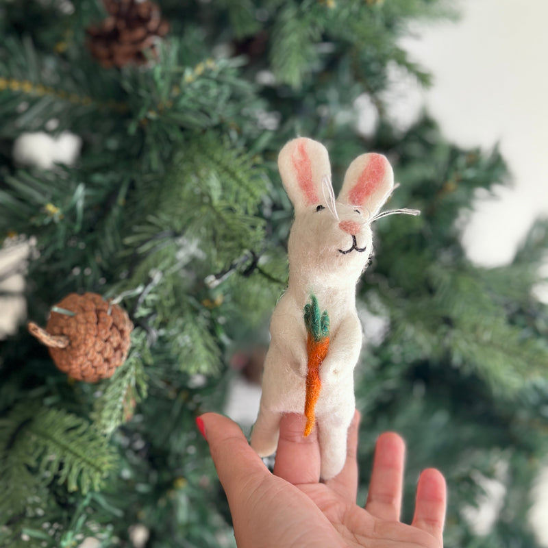 A charming felt bunny finger puppet, holding a tiny bright orange carrot, is presented in a female hand. Crafted from soft, white felt, this handcrafted puppet features long, perky ears with pink inner details and a sweet embroidered expression, evoking warmth and creative play.