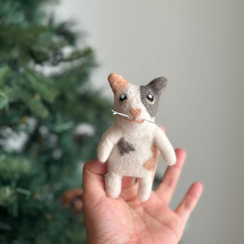 A charming needle felted calico cat finger puppet is gently held in a female hand, set against a soft, neutral background with a hint of a Christmas tree, creating a cozy, playful atmosphere. This handcrafted felt kids toy features distinct patches of peach, white, and grey, including a love spot on its round body. It has tiny arms, perky upright ears, and a unique heart-shaped pink nose.
