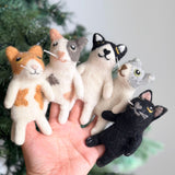 A unique handmade felt Calico Cat finger puppet is held by a female hand, appearing against a blurred background suggesting a festive Christmas tree. This charming felt toy for kids features the distinctive multi-colored fur of a calico cat, handcrafted with care.