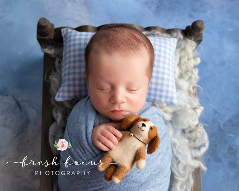 A handmade wool felt Cavalier King Charles Spaniel dog finger puppet is gently held by a baby, nestled in a miniature wood bed against a soft blue backdrop. This charming felt finger puppet features a tan body, fluffy brown ears, a sweet smile, and a tiny bell necklace, adding a whimsical and heartwarming touch to the serene scene.