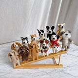 A handcrafted wool felt Cavalier King Charles Spaniel dog finger puppet sits on a small light-colored wooden stand, positioned against a cheerful backdrop of a white curtain and marble-patterned surface. This unique finger puppet, meticulously made with needle felt details, features a captivating smile, fluffy ears, an adorable love spot, and a charming bell necklace.