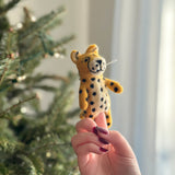A detailed, handmade, needle-felted cheetah finger puppet, crafted from 100% wool, is held up by a female hand with red nail polish. The yellow puppet features distinct black spots, rounded ears, and a small smiling mouth. Its intricate craftsmanship is highlighted as the hand provides scale against a blurred background of a Christmas tree and a window, conveying a warm, whimsical scene.