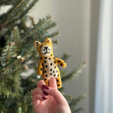A handcrafted wool felt cheetah finger puppet is presented on a finger with dark red nail polish, positioned against the soft glow of a Christmas tree. This unique needle-felted puppet, made from 100% wool, features a vibrant yellow body with distinct black spots, rounded ears, and a detailed face complete with whiskers.