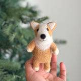 A handcrafted wool felt corgi finger puppet is held playfully in a female hand against a blurred green Christmas tree background. The adorable light brown corgi features small white feet and a distinctive white and tan face with perky ears and a small black nose, emphasizing its unique design and tactile wool felt texture.