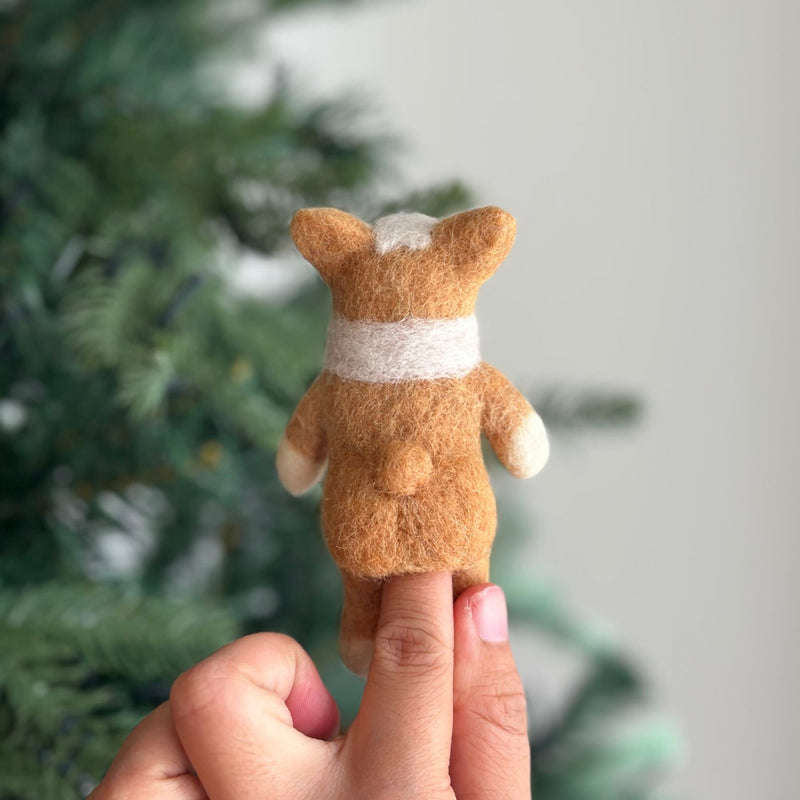 A charming handmade felt corgi finger puppet is held upright on a hand, positioned in front of a blurred Christmas tree, highlighting its festive appeal. This delightful felt toy for kids is showcased from the back, revealing its light brown fur, short limbs, and distinctive exaggerated puffy butt. The puppet features a white marking on its head, white paws, and a white band wrapped around its body.
