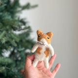 A handmade white and orange wool felt Ginger Cat finger puppet is held gently in a female hand, presented against a soft, blurred Christmas tree background. This charming felt finger puppet features an orange patch on its face, a distinctive love-shaped accent on its body, upright ears, black eyes, a stitched black mouth, and delicate white whiskers, perfect for imaginative play.