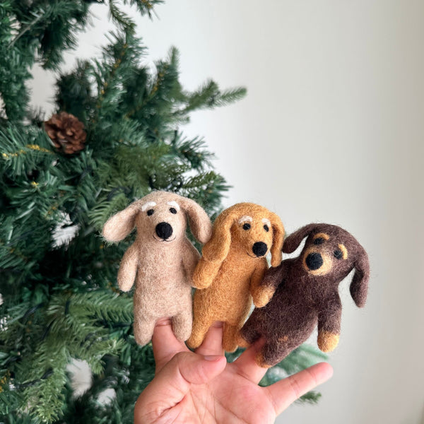 Three felt Dachshund dog finger puppets, in beige, khaki, and brown, are shown held in a female hand. A festive Christmas pine tree is blurred in the background. Each unique felted puppet beautifully captures the Dachshund's distinctive elongated body and floppy ears, making them ideal for engaging storytelling and imaginative play.