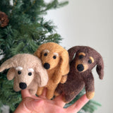 Three felt dachshund finger puppets are held in a female hand against a festive Christmas tree backdrop. These handmade felted finger puppets come in distinct beige, khaki, and brown shades, each capturing the dachshund's elongated body. Each charming felt dog features round black eyes, noses, and tiny felted eyebrows for expressive play.