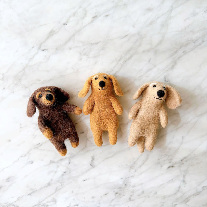 Three charming felt dachshund finger puppets are playfully arranged on a white marble surface. These handcrafted felt dog puppets come in distinct brown, khaki, and beige colors, each featuring the breed's elongated body, floppy ears, and a small stitched black nose. The brown dachshund has tan paws and ears, and the beige one displays white markings above its eyes.