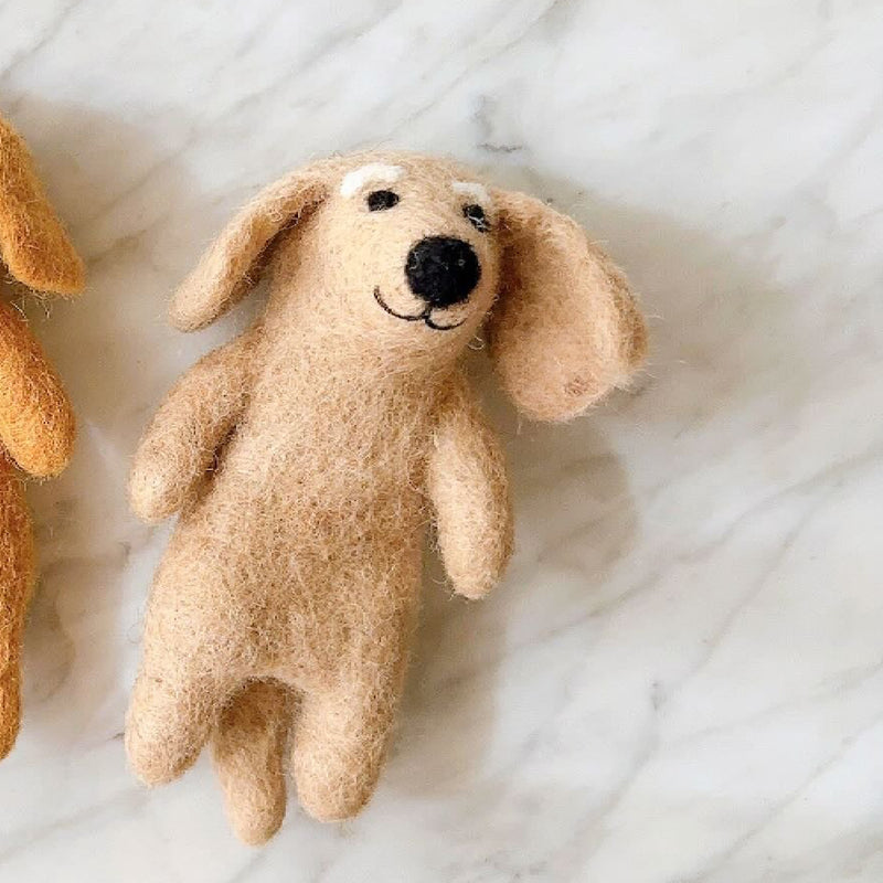 A charming, handcrafted beige dachshund felt finger puppet rests on a bright marble surface. This unique finger puppet features khaki and brown accents, embodying the breed's elongated body and floppy ears. Its soft wool felt texture, a hallmark of fair-trade craftsmanship, includes a small black nose and an embroidered smile, enhancing its playful and endearing appearance. Another similar felt finger puppet is partially visible in the background.