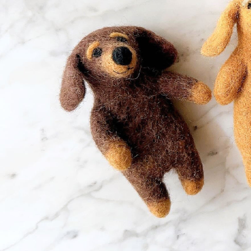 Two handmade felt dachshund finger puppets are displayed on a bright white marble surface. One brown dachshund puppet features an elongated body with beige paws, ears, and eyebrows. It rests beside another felt dachshund finger puppet. These fair-trade puppets, handcrafted in Nepal, capture the breed's charming personality.