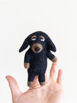 A handcrafted felt Dachshund finger puppet is held in a female hand against a bright white backdrop. This charming puppet highlights the Dachshund's distinctive elongated form, featuring small, pointed ears and minimalist facial features.