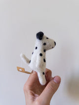 A handmade felt dalmatian dog finger puppet is held up by a female hand against a light background. This unique felt puppet features a white body with characteristic black spots, black ears, and a small attached tag.