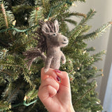 A handcrafted wool felt donkey finger puppet is held by a female hand against a festive Christmas tree adorned with white lights. This playful gray donkey finger puppet features a darker mane and tail, a light gray face, and stitched facial details, showcasing its unique design.