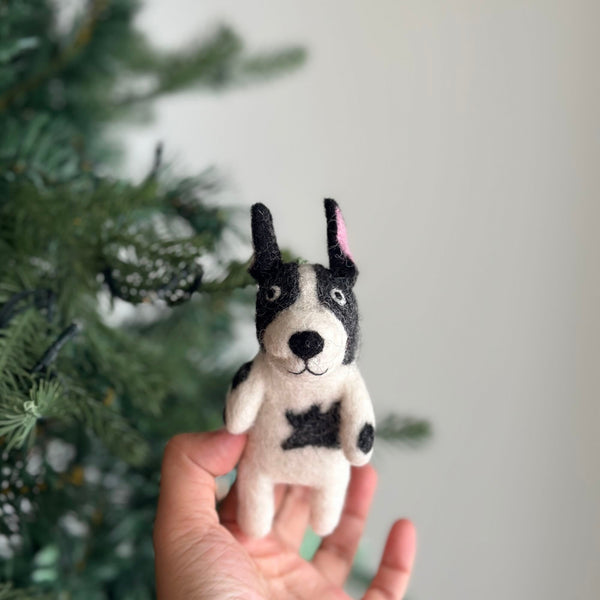 A handcrafted wool felt French bulldog finger puppet is playfully held by a female hand, with a blurred Christmas tree in the background. This unique felt toy for kids is white with distinct black markings, including a body spot, large black ears with one pink inside, and a stitched smile, showcasing meticulous needle-felted craftsmanship.