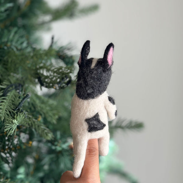 A handcrafted wool felt French bulldog finger puppet is displayed on a female hand, set against a blurred Christmas tree backdrop. This charming black and white puppet features a black head with large, pink-lined erect ears and a compact white body with a black marking on its rear. Meticulously crafted from wool felt, this fair-trade felt toy for kids sits on the fingertip, ready for engaging imaginative play.