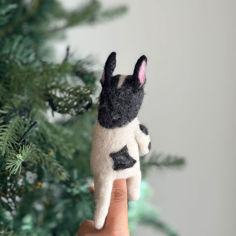 A handcrafted wool felt French bulldog finger puppet is displayed on a female hand, set against a blurred Christmas tree backdrop. This charming black and white puppet features a black head with large, pink-lined erect ears and a compact white body with a black marking on its rear. Meticulously crafted from wool felt, this fair-trade felt toy for kids sits on the fingertip, ready for engaging imaginative play.