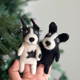 Two handcrafted wool felt French bulldog finger puppets are held in a hand against a blurred Christmas tree, suggesting a cozy, holiday setting. Meticulously crafted with needle felt detailing, one puppet is white with black markings, and the other is black with white facial details. Both feature the French Bulldog's distinct 'bat ears' for engaging, imaginative play.