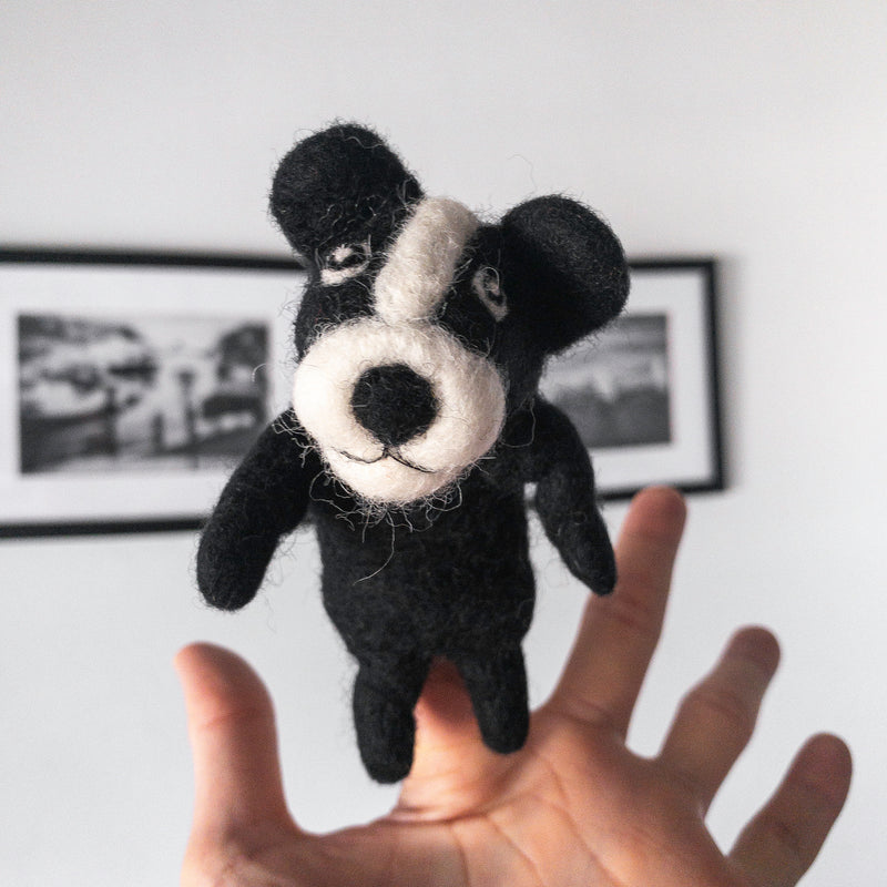 A handmade wool felt French bulldog finger puppet is presented by a female hand against a bright, minimalist background. This black and white puppet is meticulously crafted with needle felt detailing, featuring large, erect 'bat ears,' a white head patch, and a white snout. The charming puppet is held, ready for imaginative play.