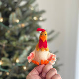 A handmade felt rooster finger puppet is held by a hand, set against a blurred Christmas tree background with soft lights. This bright and colorful felt rooster puppet features a yellow head, a vibrant red comb and wattle, and an orange beak. Its light cream body is detailed with hints of orange on the wings and feet, creating a charming, playful design.