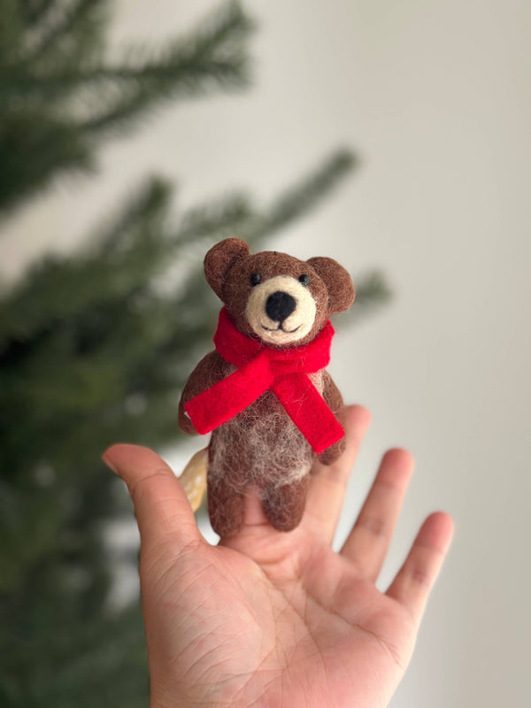 A handcrafted felt honey bear finger puppet is held upright in an open hand, with a blurred Christmas tree in the background. This unique, fair-trade honey bear puppet is crafted from brown and light gray felt, featuring a round head, small ears, and a light gray muzzle with a black nose. A bright red scarf is tied around its neck, adding a festive touch.