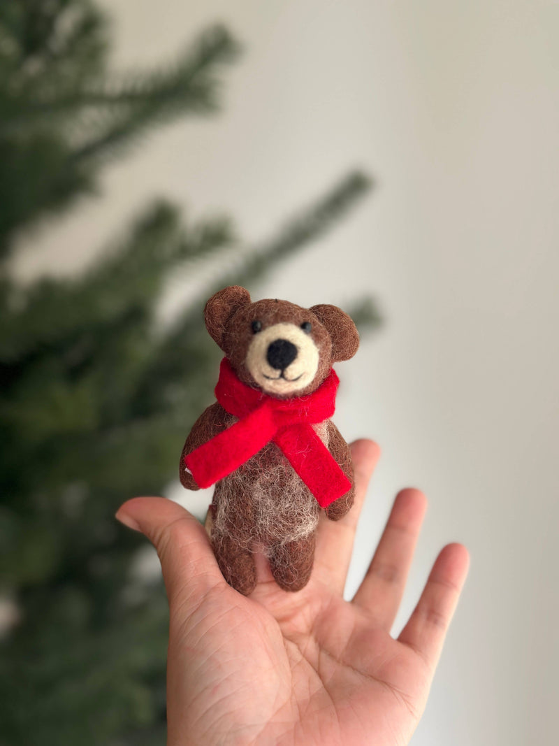 A felt Honey Bear finger puppet is held gently in a female hand, presented in a cozy close-up view with a soft, festive background hinting at a Christmas tree. This handmade wool felt Honey Bear has a brown body, rounded ears, a light muzzle, a dark nose, and a cheerful red scarf. Its soft, tactile texture highlights the endearing craftsmanship, making it a unique felt toy for kids, crafted with fair-trade principles.