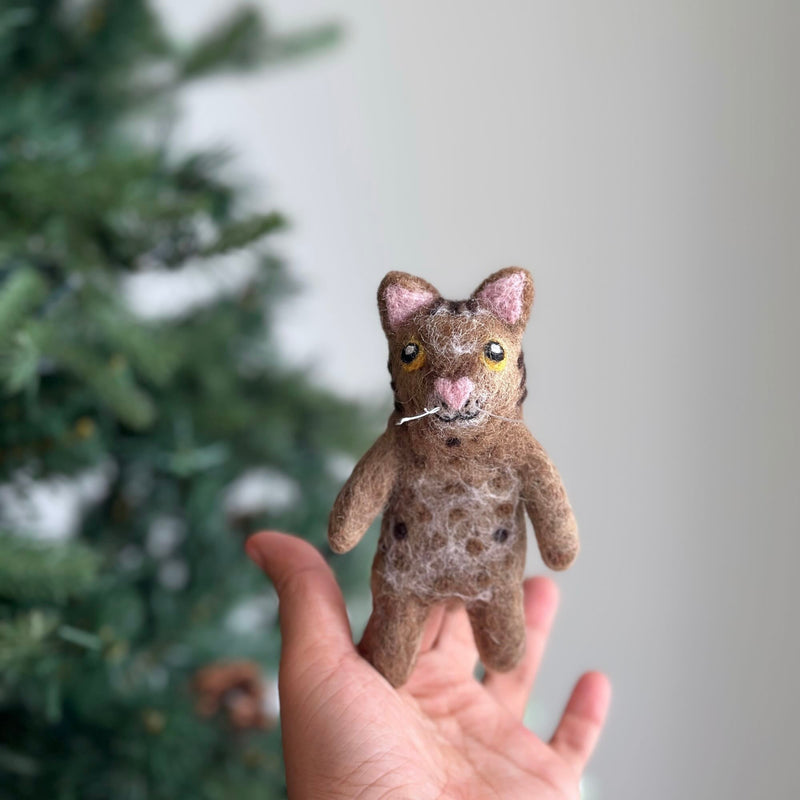 A charming, handcrafted wool felt brown tabby cat finger puppet is held delicately upright in a female hand, set against a soft, light backdrop near a Christmas tree. This felt tabby cat features a brown body with dark spots, a light pink love-shaped nose, and perky ears lined with pink felt, showcasing its expressive yellow eyes and detailed texture.