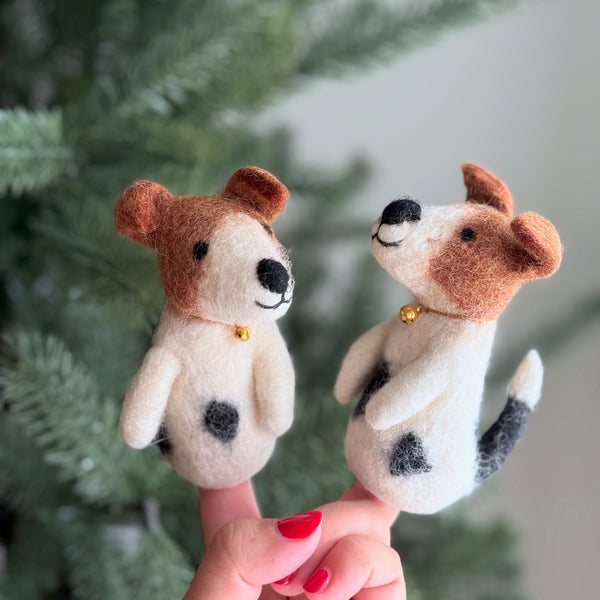 Two handmade wool felt Jack Russell Terrier finger puppets are held on a female hand, set against a soft-focus Christmas pine tree. Each brown and white needle-felted Jack Russell Terrier finger puppet features intricate details, a golden bell necklace, folding ears, an adorable love spot, and an irresistible stitched smile. One puppet looks to the right, while the other faces straight ahead, capturing their playful charm.