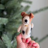 A handmade wool felt Jack Russell Terrier dog finger puppet is delicately held by a female hand with red nail polish, set against a blurred festive background with a Christmas pine tree. This unique needle-felted puppet is predominantly white with brown markings on its head and folded ears. It features an adorable black love spot, an irresistible smile, and a golden bell necklace, showcasing exquisite craftsmanship.