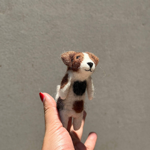 A handmade wool felt Jack Russell Terrier dog finger puppet is held by a hand with red nail polish, presented against a muted grey background. This unique finger puppet is crafted from white wool felt, featuring distinct brown and black spots, small folding ears, and a black stitched smile.