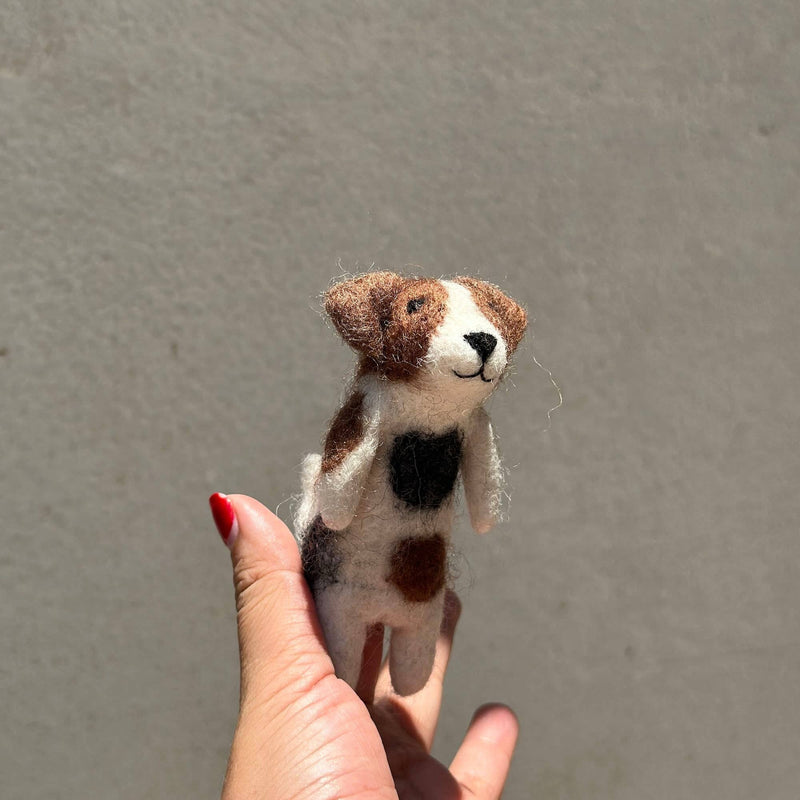 A handmade wool felt Jack Russell Terrier dog finger puppet is held by a hand with red nail polish, presented against a muted grey background. This unique finger puppet is crafted from white wool felt, featuring distinct brown and black spots, small folding ears, and a black stitched smile.