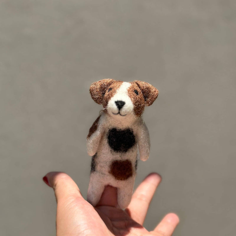 A female hand holds a handcrafted wool felt Jack Russell Terrier dog finger puppet, featuring meticulous needle-felt details. This unique finger puppet has brown and white mottled fur, dark brown folding ears, and an adorable love spot on its chest. It wears a bell necklace and sports an irresistible smile, capturing a playful and friendly essence.