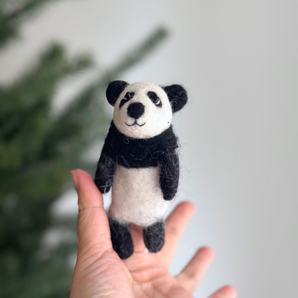 A handmade felt Kung Fu Panda finger puppet is gently held by a female hand, set against a softly blurred Christmas tree background. This charming handcrafted felt kids toy features a predominantly white body with black accents around its eyes, ears, arms, and feet, echoing the iconic character's design. Its soft texture invites imaginative play.