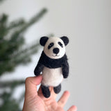 A handmade felt Kung Fu Panda finger puppet is gently held by a female hand, presented against a soft, out-of-focus background featuring a glimpse of a Christmas tree branch. This unique felt panda puppet showcases its signature black ears, eye patches, and limbs, sharply contrasting with its white body. Its detailed craftsmanship and soft texture invite a playful and tactile experience.
